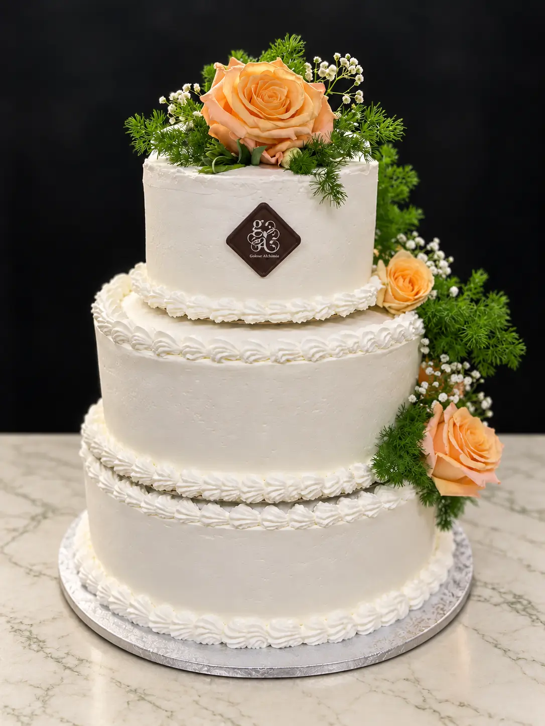 Torta Golose Alchimie a tre piani color avorio, decorata con rose color pesca, su un piano in marmo chiaro con venature salvia